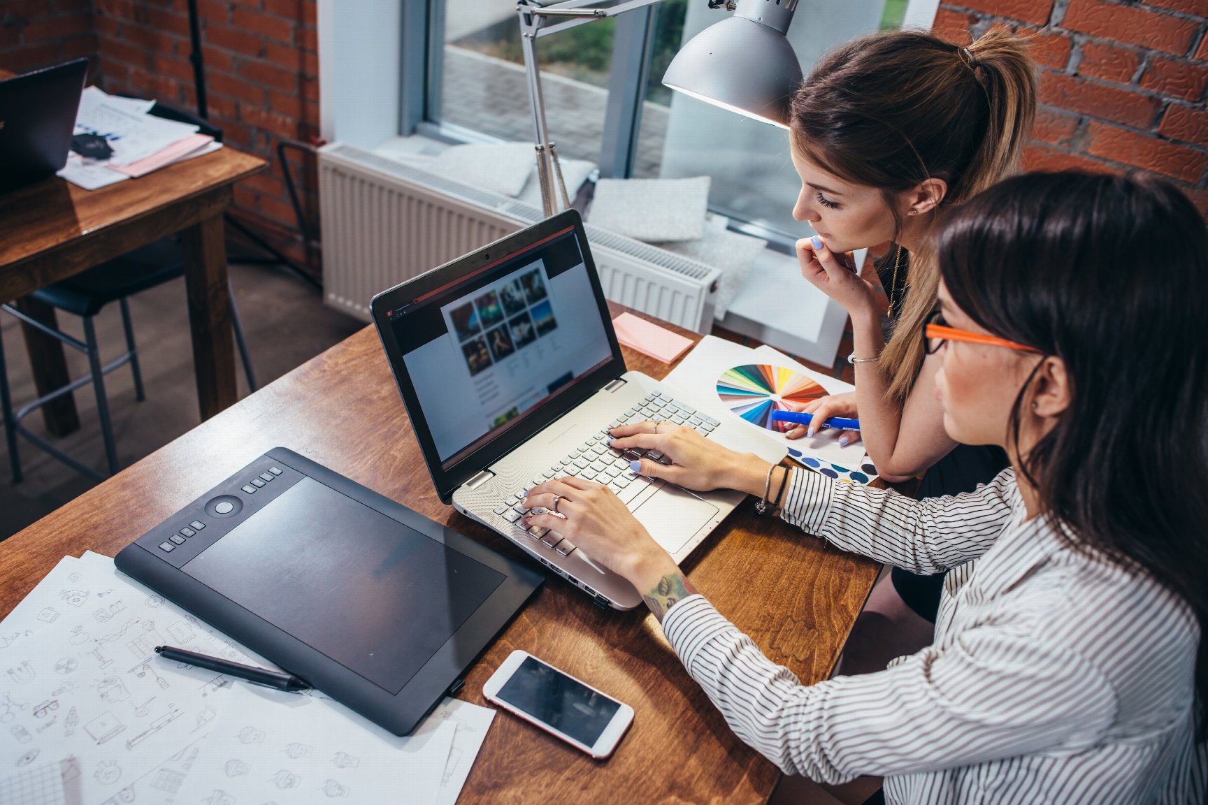 Deux femmes travaillent sur un ordinateur portable, entre une tablette graphique et un nuancier de couleurs